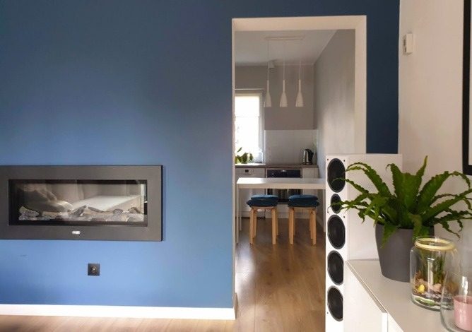 Salon z kominkiem elektrycznym w zabudowie, widok na kuchnię przez otwarte przejście, drewniana podłoga, białe głośniki i roślina doniczkowa na szafce.