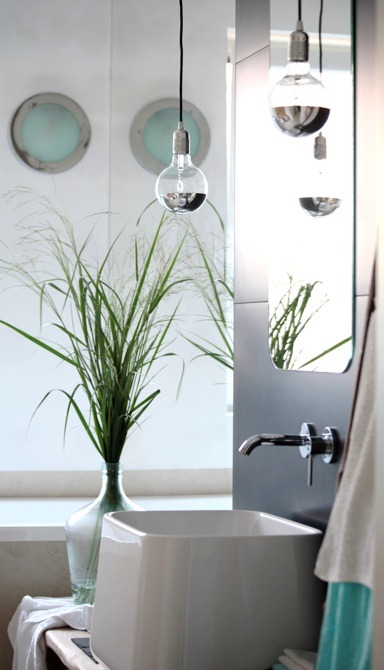 Nowoczesna łazienka z białą, prostokątną umywalką, chromowaną baterią, wiszącymi żarówkami dekoracyjnymi i trawą w szklanym wazonie na tle okrągłych okien.