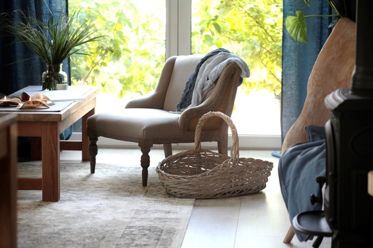 Beżowy fotel z poduszką i pledem w jasnym wnętrzu, wiklinowy koszyk na podłodze, drewniany stolik z dekoracjami, widok na zieleń za oknem.