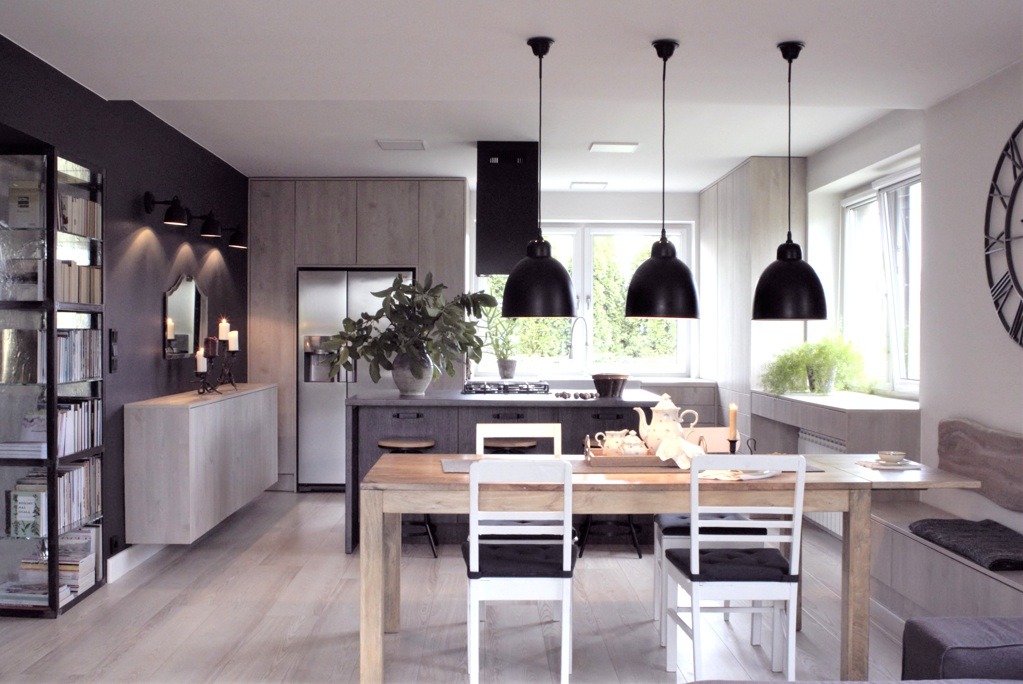 Jasna kuchnia z jadalnią w stylu skandynawskim, drewniany stół z białymi krzesłami, czarne lampy wiszące, szara zabudowa kuchenna i drewniana podłoga.