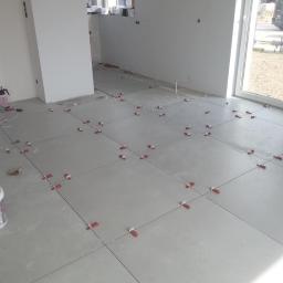 Remdom - Układanie szarych płytek podłogowych z użyciem czerwonych klinów poziomujących w jasnym pomieszczeniu z dużym oknem.