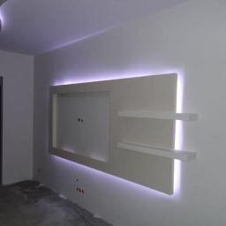 Remdom - Biała ściana z podświetlanym panelem dekoracyjnym i półkami, w surowym stanie, z widocznymi kablami elektrycznymi, przed wykończeniem podłogi.