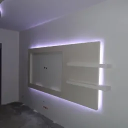 Biała ściana z podświetlanym panelem dekoracyjnym i p&oacute;łkami, w surowym stanie, z widocznymi kablami elektrycznymi, przed wykończeniem podłogi.