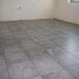 Remdom - Szare płytki podłogowe ułożone w kwadraty z białymi krzyżykami dystansowymi, widoczne ślady po kleju na niektórych płytkach, surowe ściany w tle.