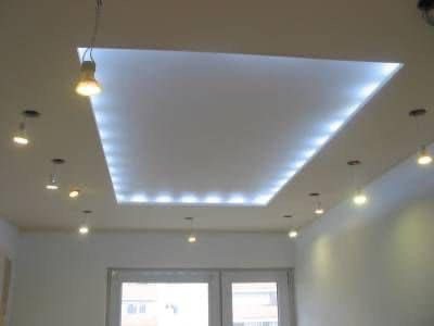 Biały sufit podwieszany z kwadratowym wnęką oświetloną diodami LED, uzupełniony o punktowe lampy halogenowe i wiszącą żarówkę, widoczny na tle białych ścian i okna.