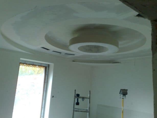 Niedokończony sufit z centralnym okrągłym elementem gipsowym i wbudowanymi otworami wentylacyjnymi, widoczne ślady szpachlowania, drabina i lampa na statywie w tle.