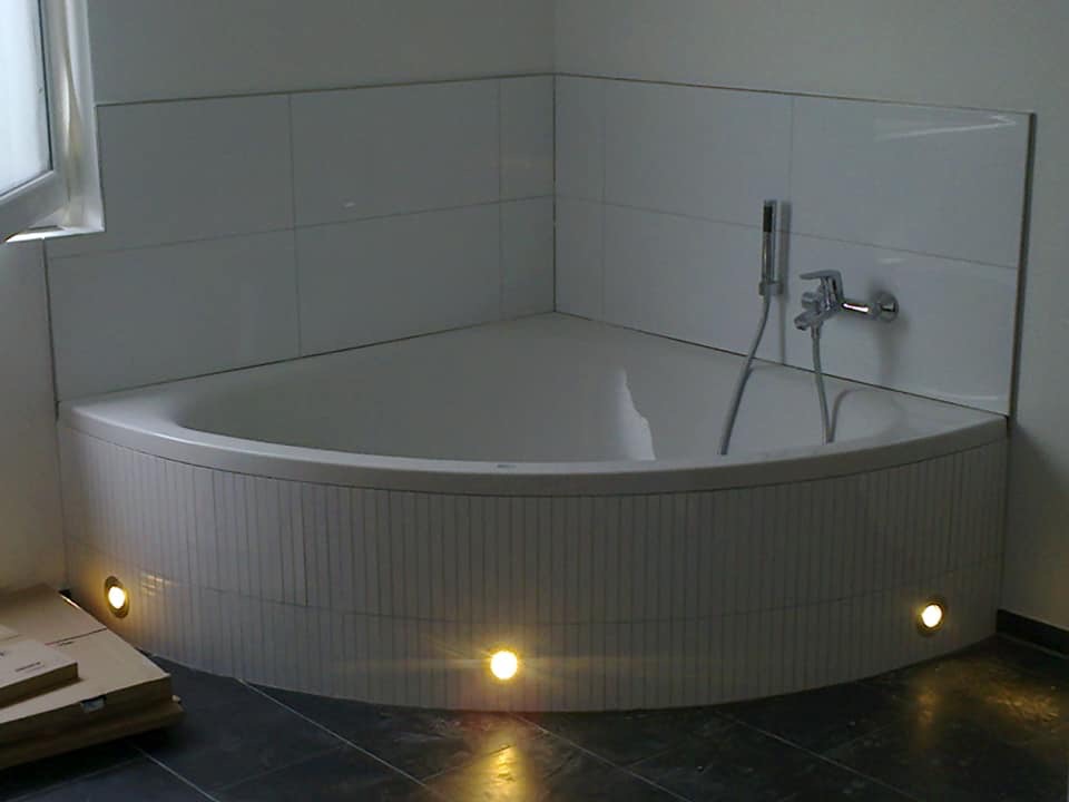 Narożna wanna z hydromasażem, obudowana mozaiką z podświetleniem LED, bateria prysznicowa na białych płytkach, ciemna podłoga.