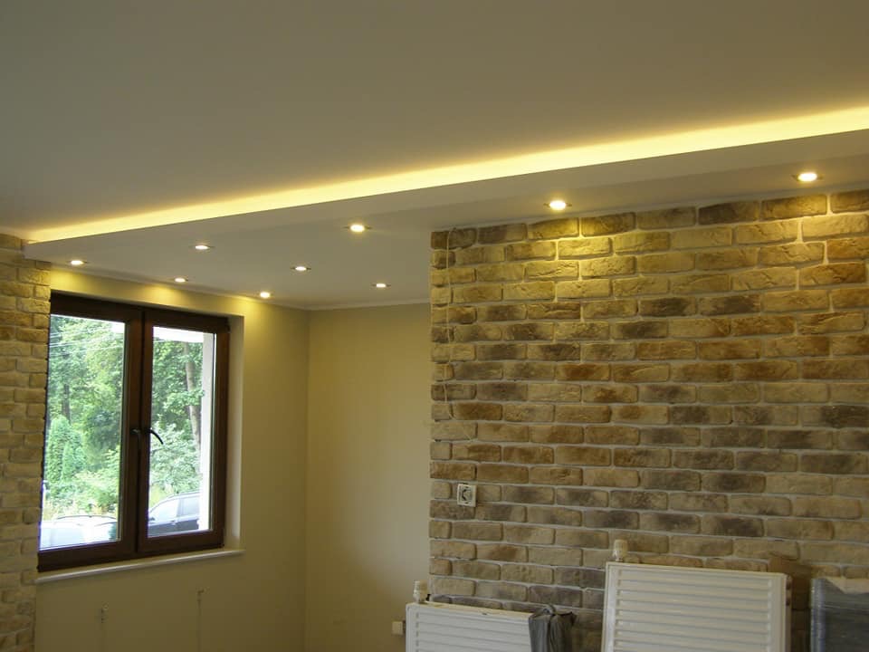 Pokój z wykończoną ścianą z cegły dekoracyjnej, podwieszanym sufitem z oświetleniem punktowym i oknem z widokiem na zieleń.