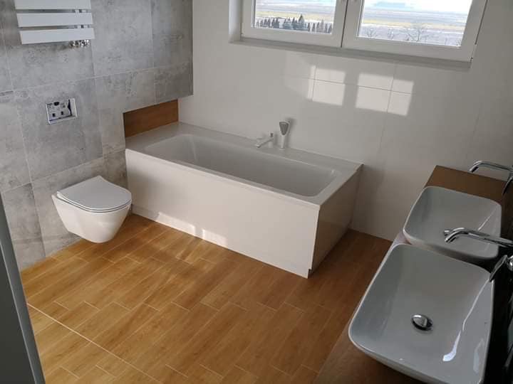 Nowoczesna łazienka z drewnopodobną podłogą, wiszącą toaletą, białą wanną i dwiema umywalkami nablatowymi na drewnianym blacie, widok na krajobraz za oknem.