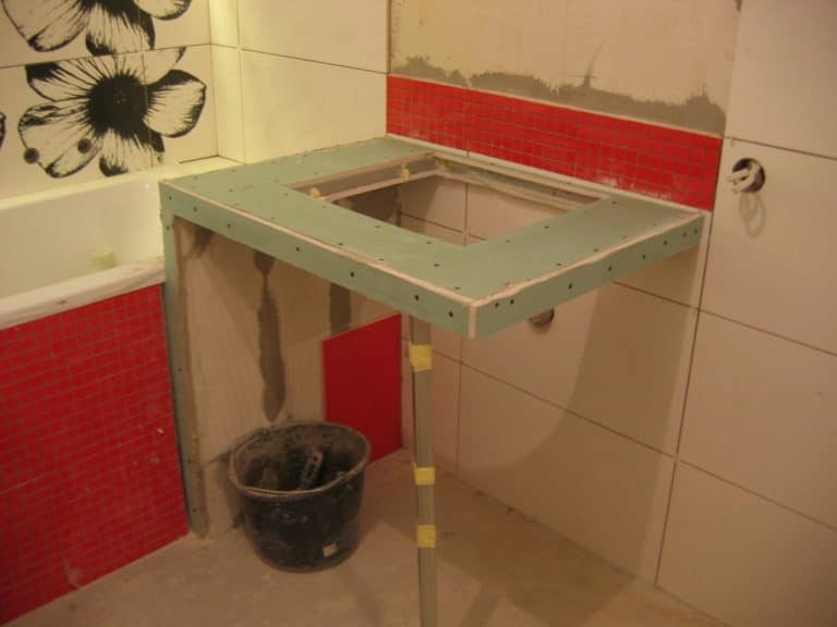 Łazienka w trakcie prac: konstrukcja pod umywalkę z płyty gipsowo-kartonowej, widoczne płytki z motywem kwiatowym i czerwona mozaika, kubeł z zaprawą na podłodze.