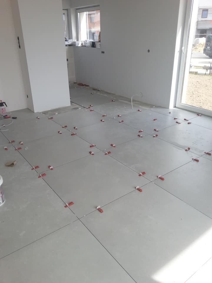 Układanie szarych płytek podłogowych z użyciem czerwonych klinów poziomujących w jasnym pomieszczeniu z dużym oknem.