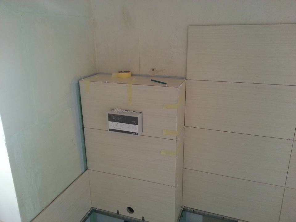 Fragment łazienki w trakcie wykańczania: obudowa stelaża WC wyłożona płytkami, zabezpieczona taśmą malarską, widoczny panel sterowania i ołówek na górze, ściany częściowo wykończone gładzią.