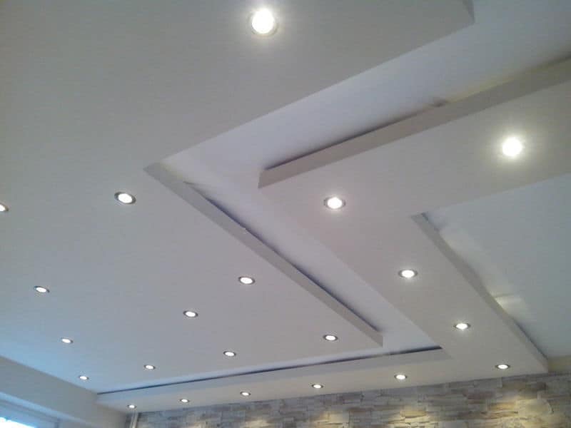 Biały sufit podwieszany z dekoracyjnymi listwami i punktowym oświetleniem LED, widoczny fragment ściany wykończonej kamieniem dekoracyjnym.