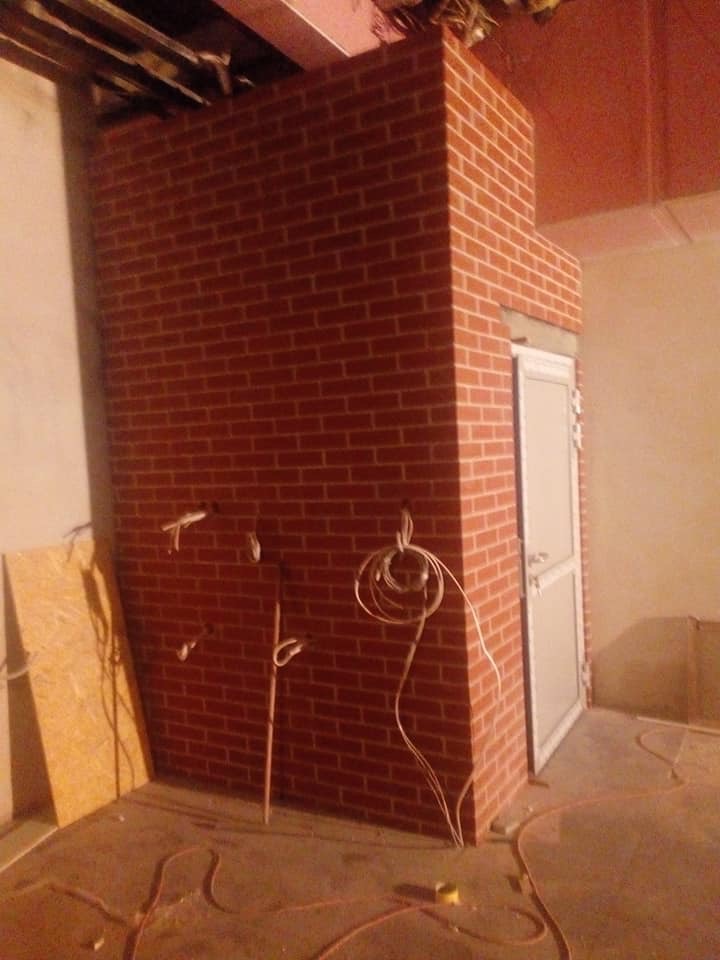 Wzniesiona ściana z czerwonej cegły z ościeżnicą drzwiową w surowym wnętrzu, widoczne przewody elektryczne i płyta OSB, w trakcie prac wykończeniowych.