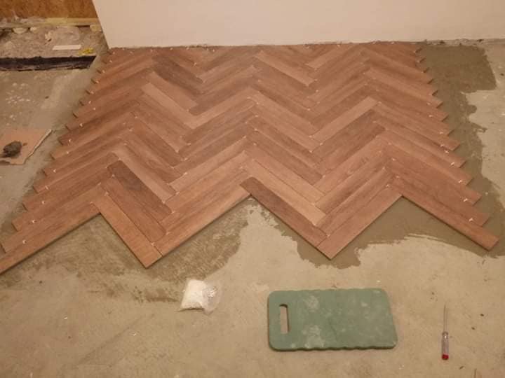 Ułożony fragment parkietu w jodełkę, widoczne plastikowe kliny dystansowe, klej i podkładka pod kolana podczas montażu.