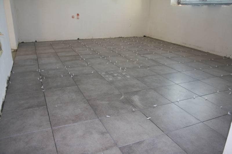 Szare płytki podłogowe ułożone w kwadraty z białymi krzyżykami dystansowymi, widoczne ślady po kleju na niektórych płytkach, surowe ściany w tle.