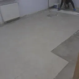 Wyrównywanie podłoża w pomieszczeniu za pomocą szpachli, widoczny pracownik w niebieskich rękawicach i wiadro z masą szpachlową, przygotowanie do dalszych prac wykończeniowych.