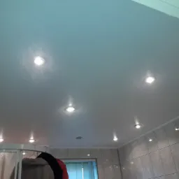 Błyszczący, jasny sufit z zamontowanymi punktowymi lampami LED, widoczne płytki ceramiczne na ścianach i fragment kabiny prysznicowej z wieszakiem na ubrania w tle.