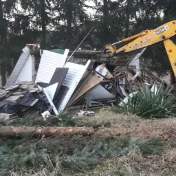 Żółta koparka JCB rozbiera zniszczony budynek z fragmentami ścian z sidingu, widoczne pozostałości dachu i wyposażenia wnętrza, w tle drzewa iglaste.