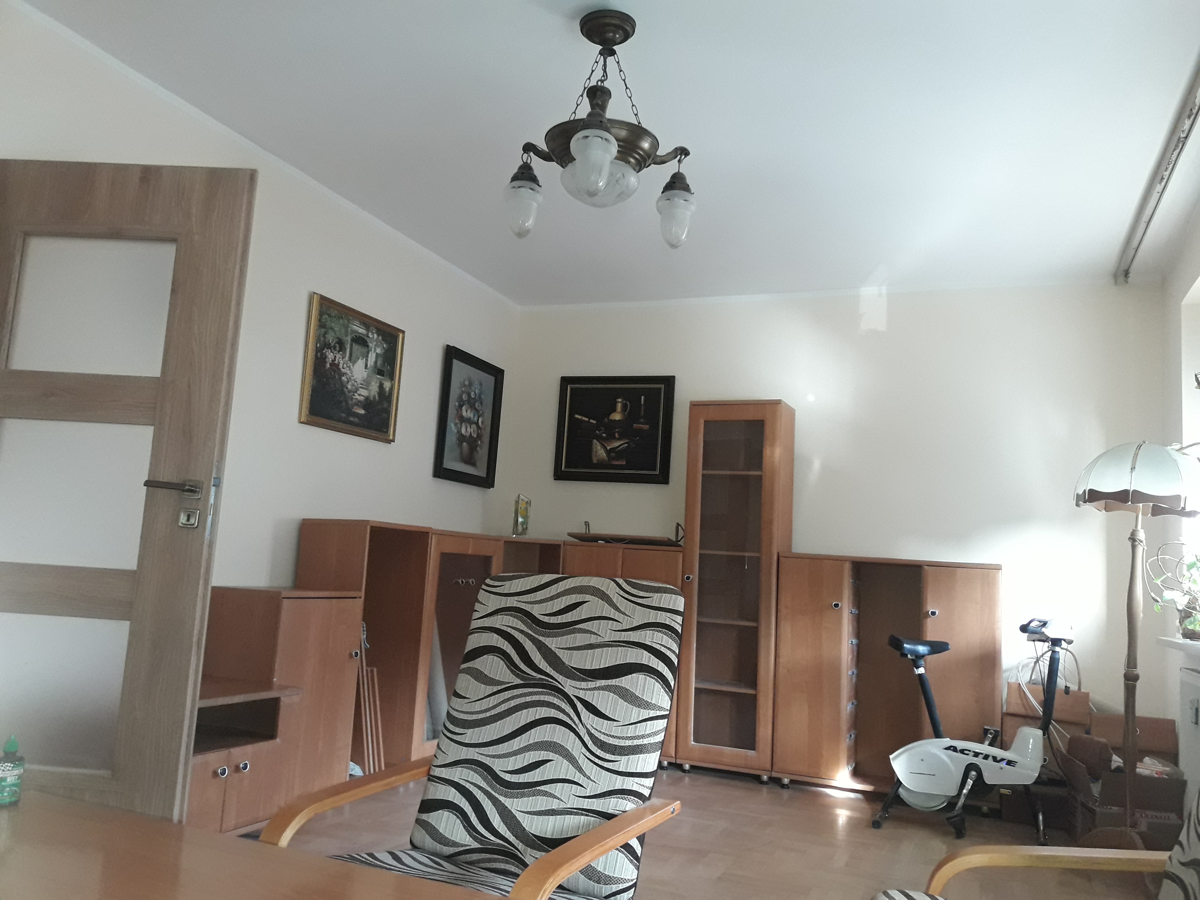 Jasne wnętrze pokoju w Kielcach z drewnianymi meblami, wzorzystym fotelem, lampą podłogową i wiszącym żyrandolem. Na ścianach wiszą obrazy.