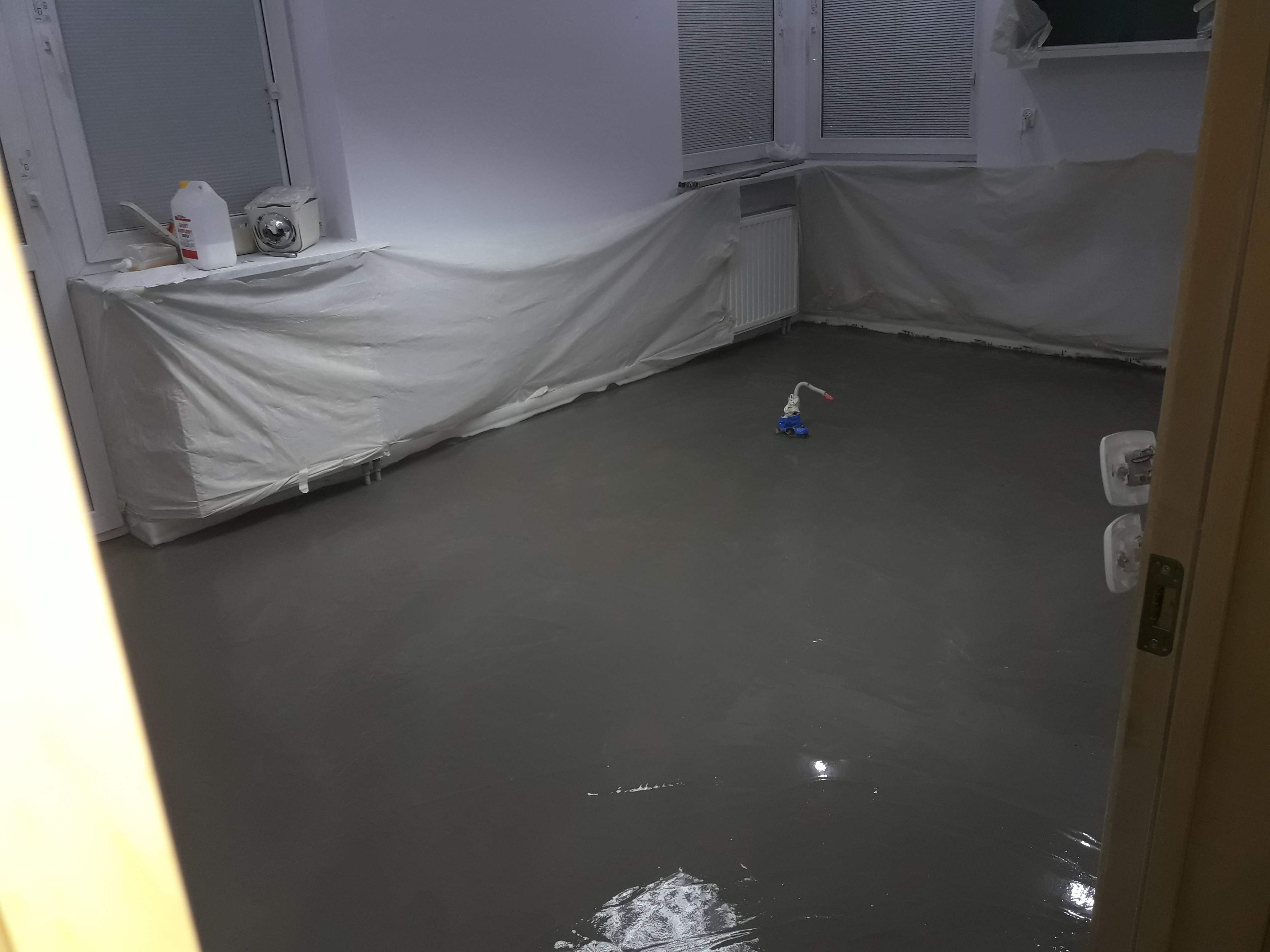 Świeżo wylana, samopoziomująca wylewka podłogowa w pokoju, zabezpieczone folią okna i grzejniki, widoczny sprzęt do rozprowadzania masy.