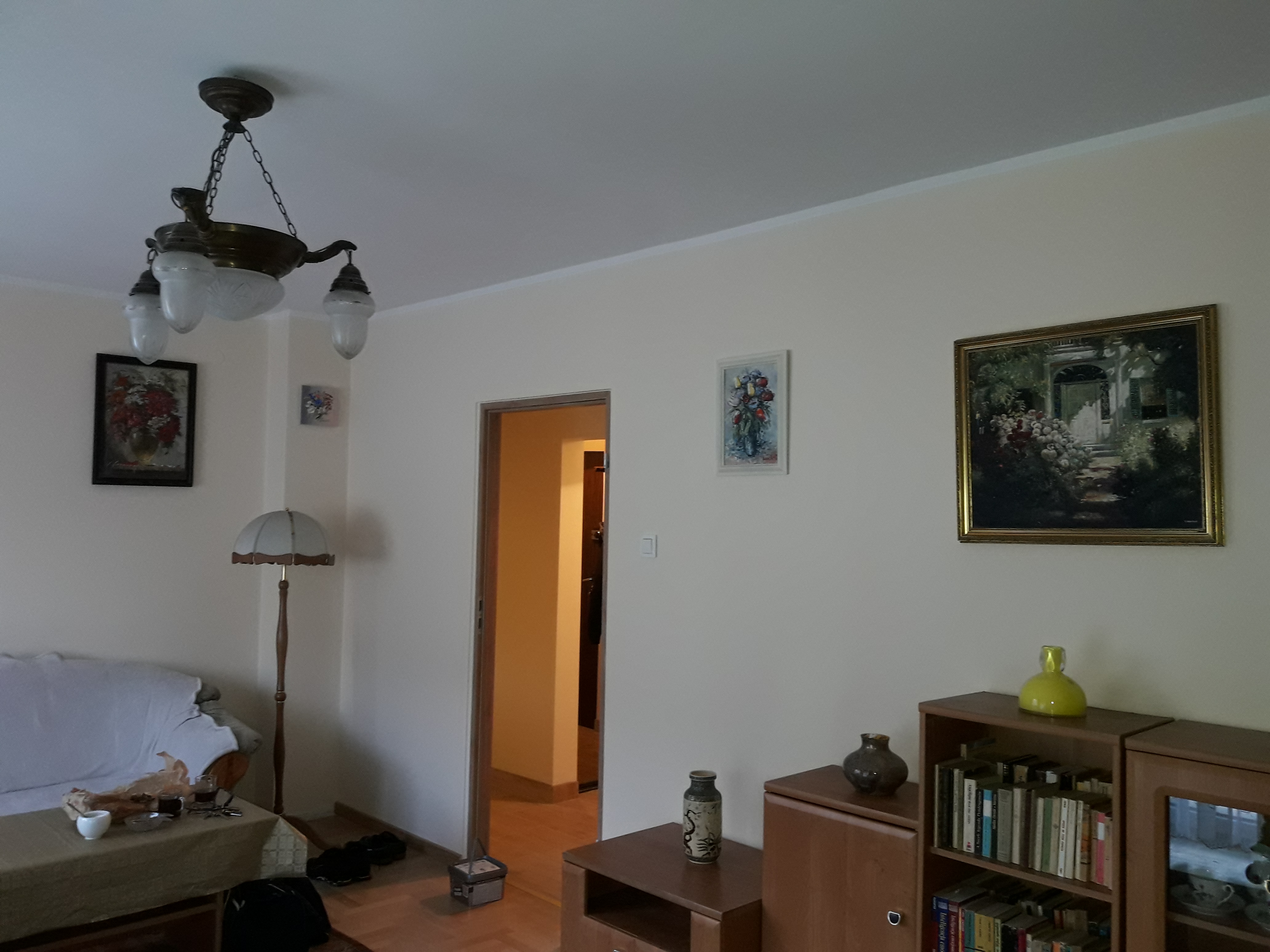 Jasne wnętrze pokoju z kremowymi ścianami, ozdobione obrazami w ramkach, z widoczną lampą wiszącą w stylu retro, stojącą lampą z abażurem, meblami w odcieniach brązu i otwartymi drzwiami...