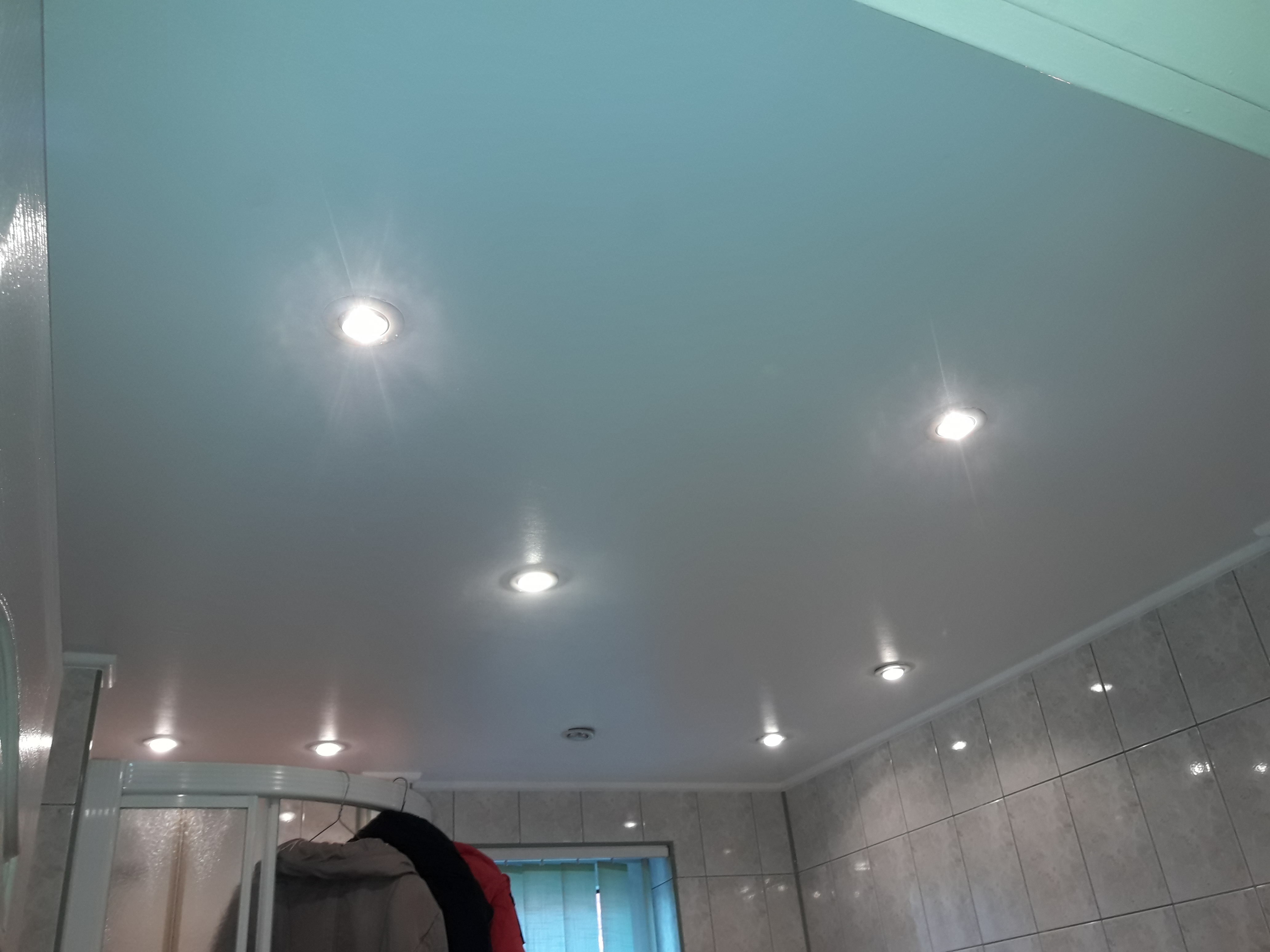 Błyszczący, jasny sufit z zamontowanymi punktowymi lampami LED, widoczne płytki ceramiczne na ścianach i fragment kabiny prysznicowej z wieszakiem na ubrania w tle.