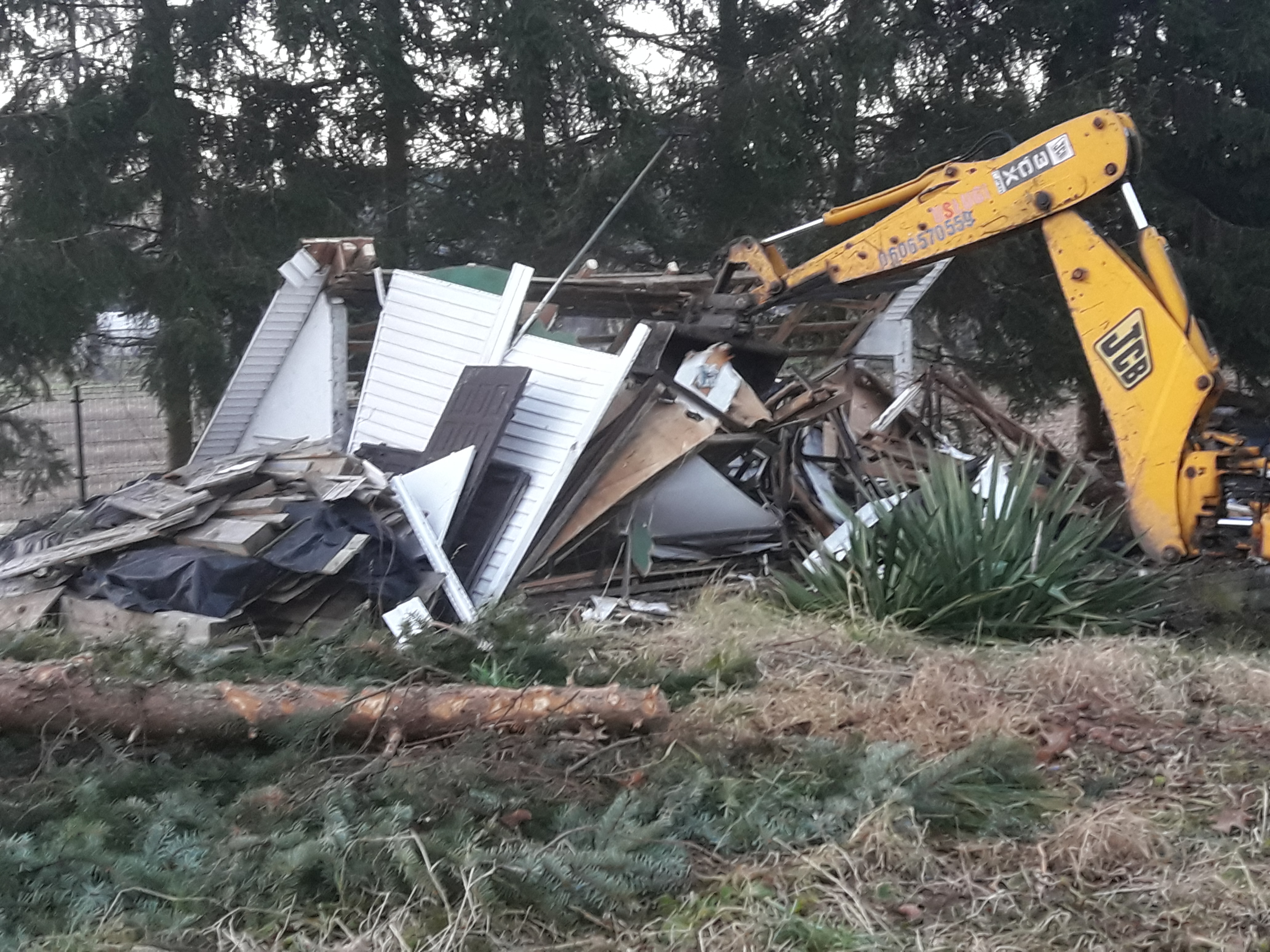 Żółta koparka JCB rozbiera zniszczony budynek z fragmentami ścian z sidingu, widoczne pozostałości dachu i wyposażenia wnętrza, w tle drzewa iglaste.