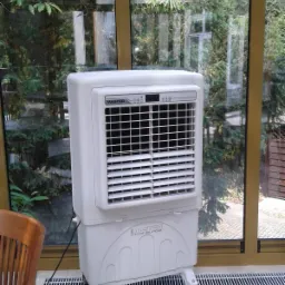 Biały przenośny klimatyzer Master na k&oacute;łkach, stojący w przeszklonym pomieszczeniu z widokiem na zieleń.