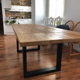 Rozkładany stół z blatem z jasnego drewna o widocznej strukturze słojów, osadzony na czarnych, metalowych nogach o prostokątnym przekroju, w tle jasna kuchnia z białymi szafkami i ceglaną ścianą...