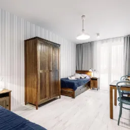 Pokój hotelowy z dwoma łóżkami, drewnianą szafą i stołem z krzesłami w stylu skandynawskim, oświetlony lampami i światłem dziennym.