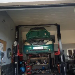 Zielony samochód z otwartą maską uniesiony na dwukolumnowym podnośniku warsztatowym w garażu z widocznymi narzędziami i wyposażeniem warsztatowym.