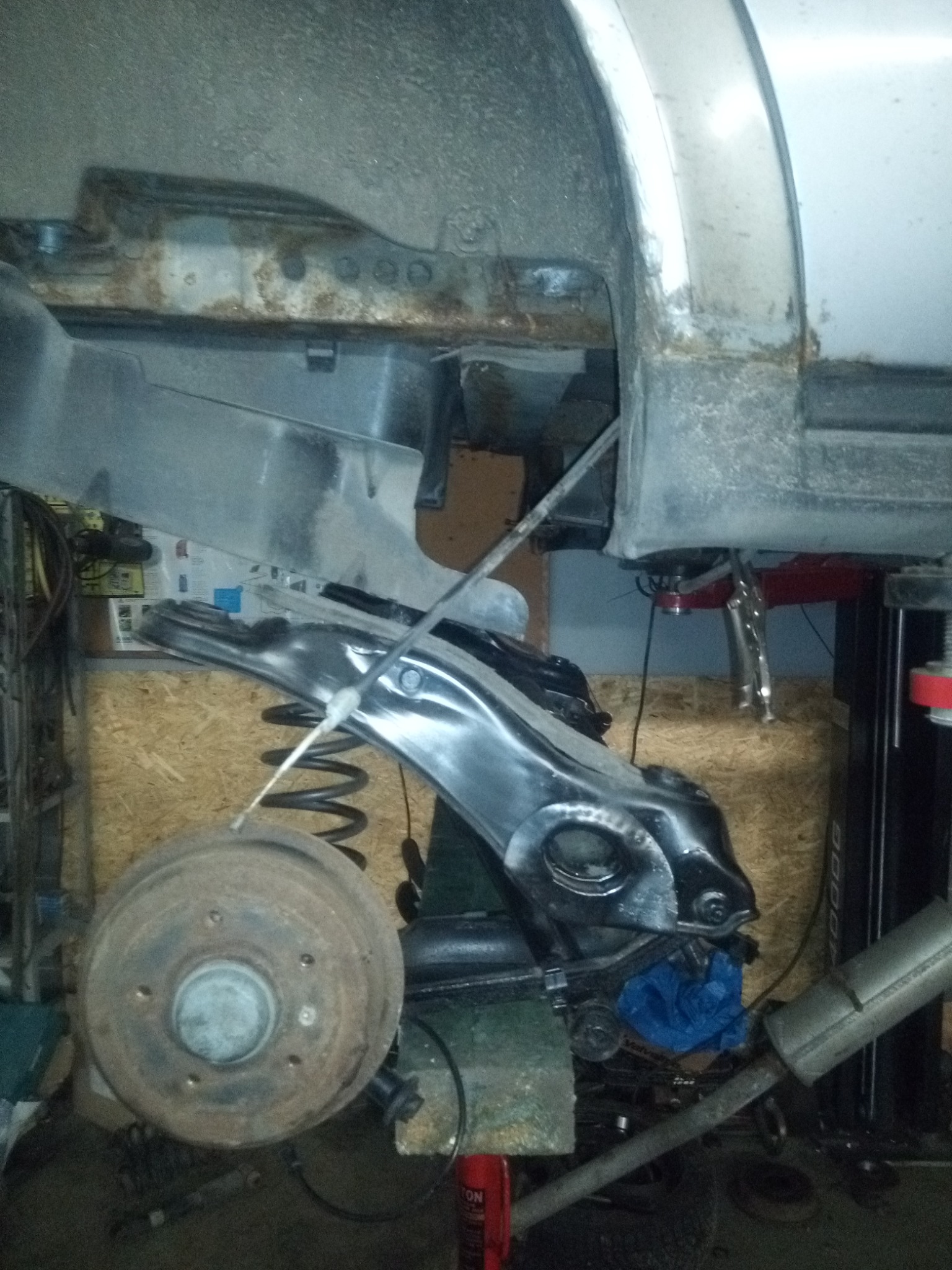 Widok podwozia samochodu na podnośniku, z widocznym skorodowanym błotnikiem, nowym wahaczem pomalowanym na czarno, sprężyną zawieszenia i bębnem hamulcowym. Elementy układu wydechowego w tle.