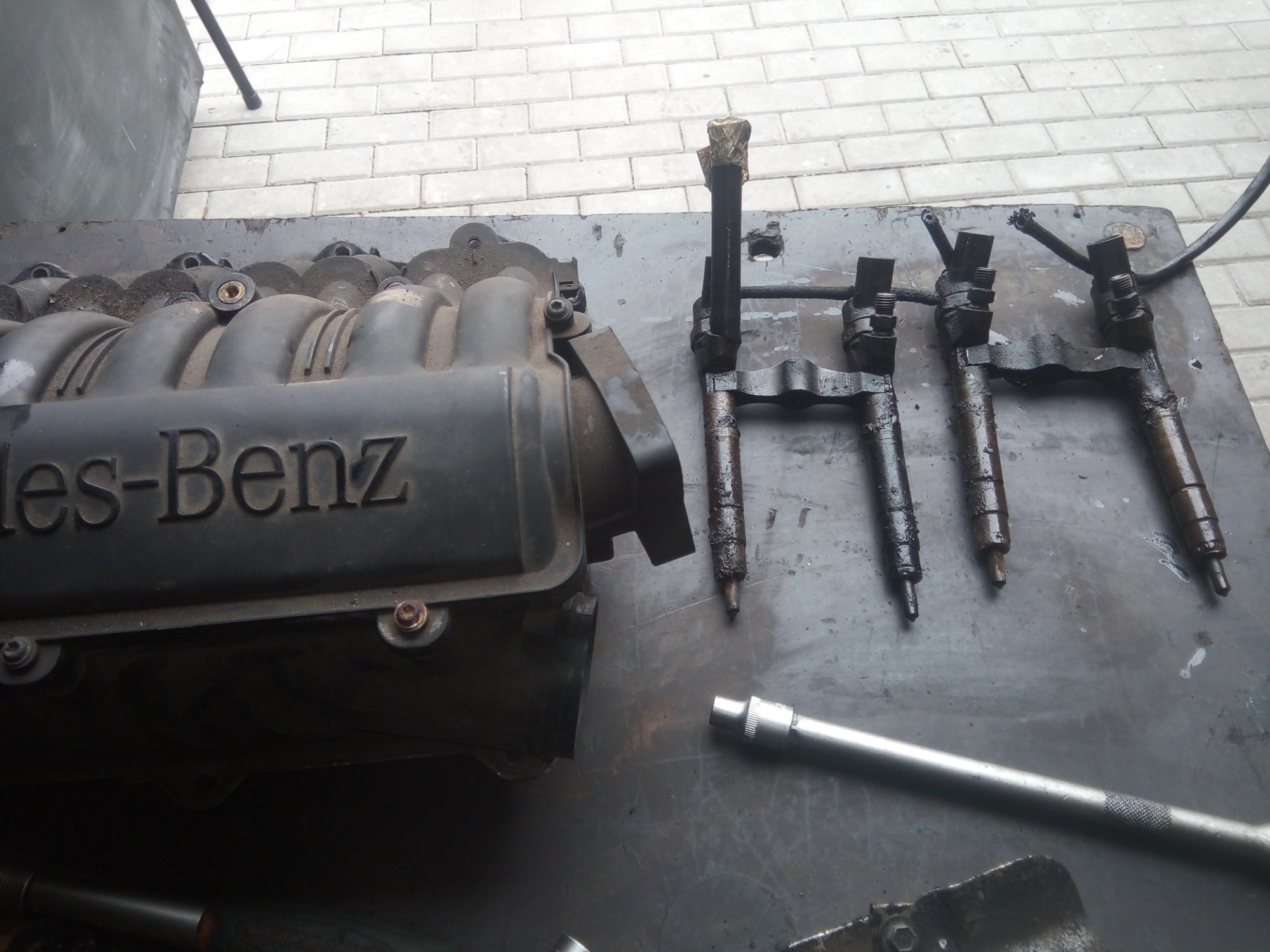 Zdemontowany kolektor dolotowy Mercedes-Benz obok wtryskiwaczy paliwa i klucza nasadowego na brudnym stole warsztatowym, widoczne ślady oleju i zabrudzenia.