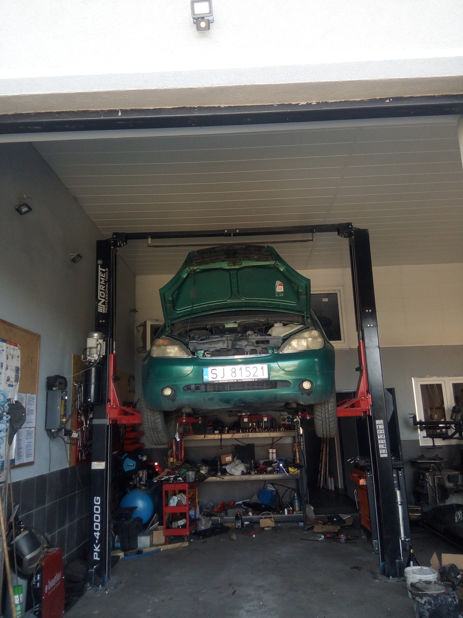 Zielony samochód z otwartą maską uniesiony na dwukolumnowym podnośniku warsztatowym w garażu z widocznymi narzędziami i wyposażeniem warsztatowym.