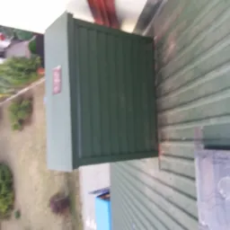 Zielona blacha trapezowa na dachu budynku, widok z góry, obok skrzynka elektryczna, w tle ogród z zielenią.