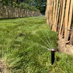 Automatyczny system nawadniania trawnika w ogrodzie, z widoczną linią drewnianego płotu i zraszaczem w akcji, rozpylającym wodę na soczystą trawę.