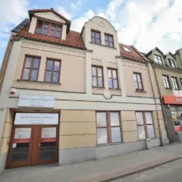 Elewacja budynku biurowego z szyldem informacyjnym przy wejściu, nad drzwiami widnieje nazwa firmy i numer telefonu.