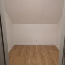 Poddasze po remoncie z nową podłogą z paneli laminowanych o ciepłym odcieniu drewna i świeżo pomalowanymi na biało ścianami, widok zza otwartych drzwi.