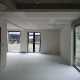 Surowe wnętrze domu w stanie deweloperskim z widocznymi gładziami na ścianach i suficie, betonowy strop z widocznymi elementami konstrukcyjnymi oraz duże okna wychodzące na ogród.