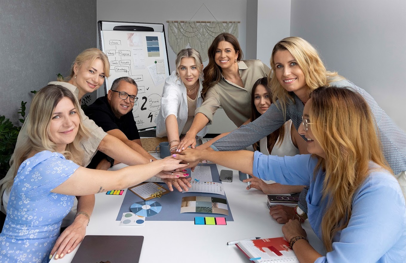 Zespół kreatywny łączy dłonie nad stołem z próbkami kolorów, notatnikiem i planem strategicznym na tablicy, demonstrując współpracę i zaangażowanie w proces tworzenia.