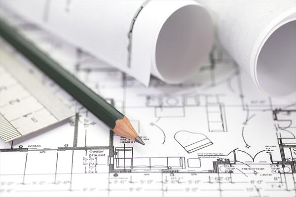 Fragment planu architektonicznego domu z naniesionymi wymiarami i elementami wyposażenia, zwinięte plany w tle, ołówek i linijka na pierwszym planie.