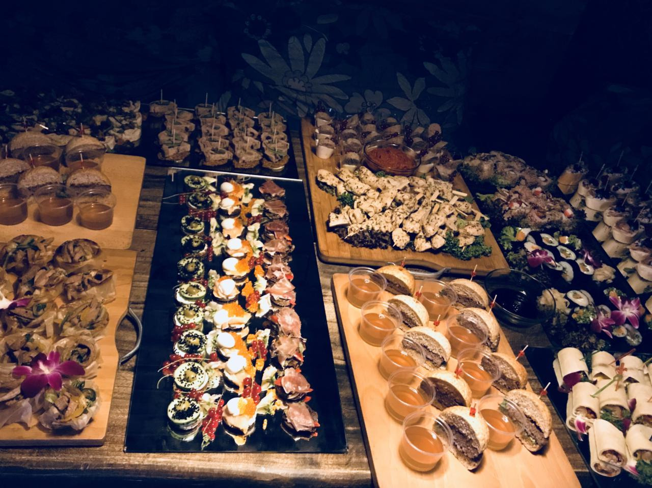 Bogato zastawiony stół z różnorodnymi przekąskami na drewnianych deskach i czarnych tacach, w tym mini burgery, szaszłyki z kurczaka, sushi, roladki i przystawki z owocami, całość w ciemnym...