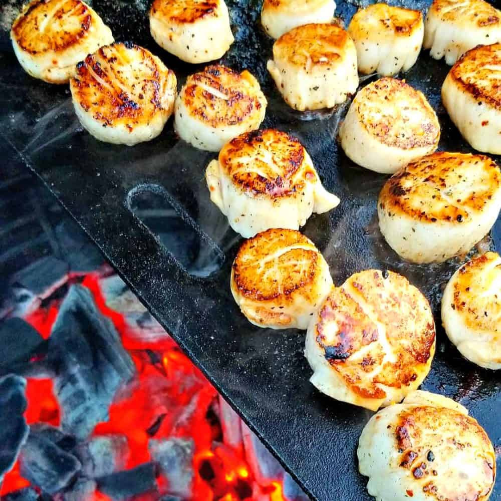 Smażone przegrzebki na czarnym grillu nad rozżarzonymi węglami, widoczne przyprawy i delikatne przypalenia na powierzchni.