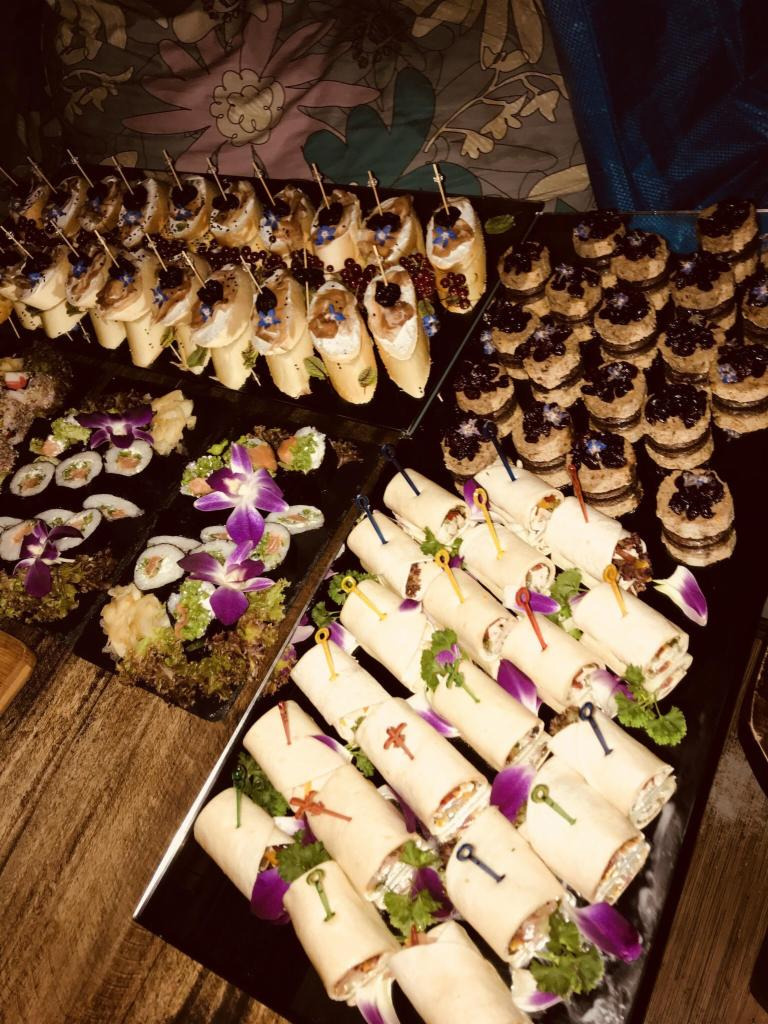 Bogaty wybór przekąsek na czarnych tacach, udekorowanych fioletowymi kwiatami, widoczny na stole w Gdańsku. Wśród nich mini tortille z kolorowymi wykałaczkami, sushi oraz kanapeczki z owocami.