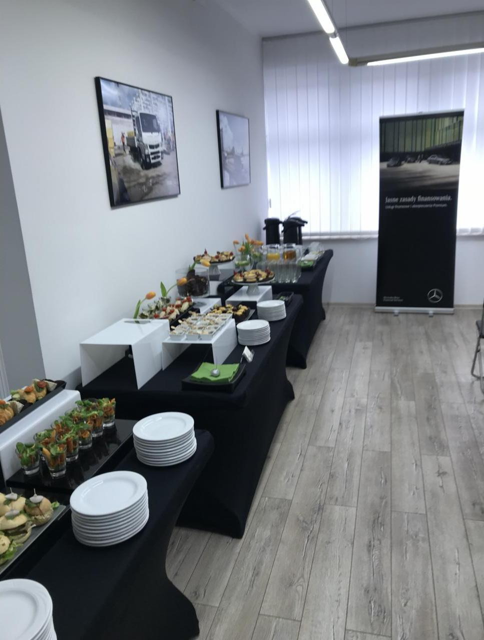 Długi stół cateringowy z czarnym obrusem, zastawiony różnorodnymi przekąskami, deserami i napojami, w tle widoczny baner reklamowy Mercedes-Benz i zdjęcia ciężarówek na ścianie, w jasnym...