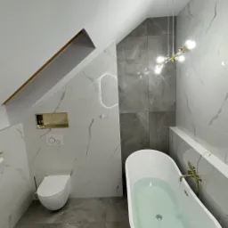 Nowoczesna łazienka na poddaszu z białą wolnostojącą wanną wypełnioną wodą, marmurowe płytki na ścianach, złote akcenty w oświetleniu i armaturze, oraz toaleta w zabudowie.