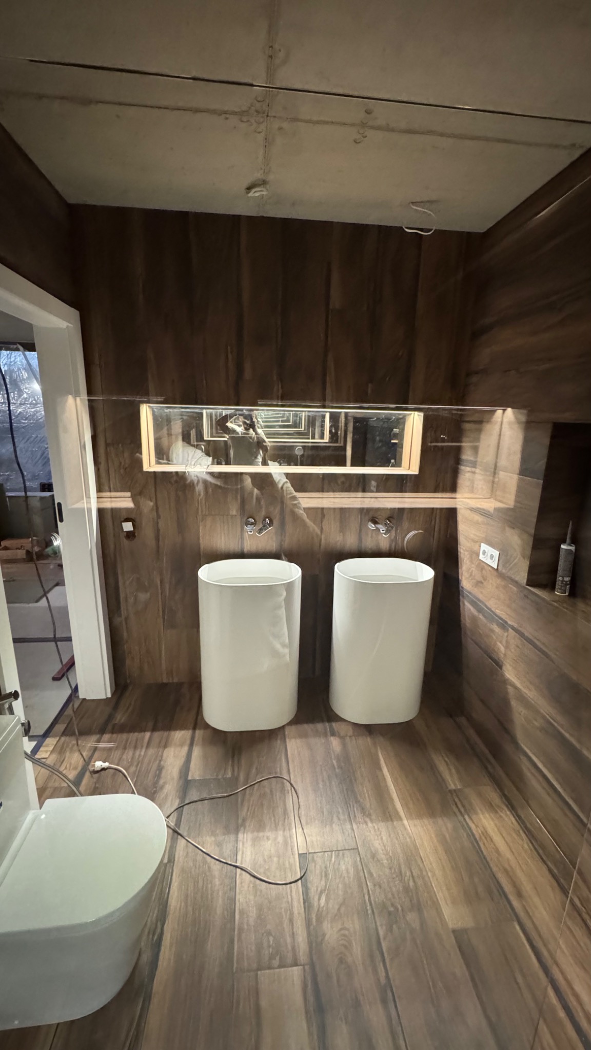 Nowoczesna łazienka z dwoma białymi umywalkami cylindrycznymi, lustrem z efektem nieskończoności i drewnopodobnymi płytkami na ścianach i podłodze. Widoczny fragment toalety.