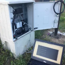 Otwarta szafka telekomunikacyjna z widocznymi kablami i urządzeniami, obok niej laptop Dell z uruchomionym terminalem diagnostycznym, otoczenie to trawa i ogrodzenie.