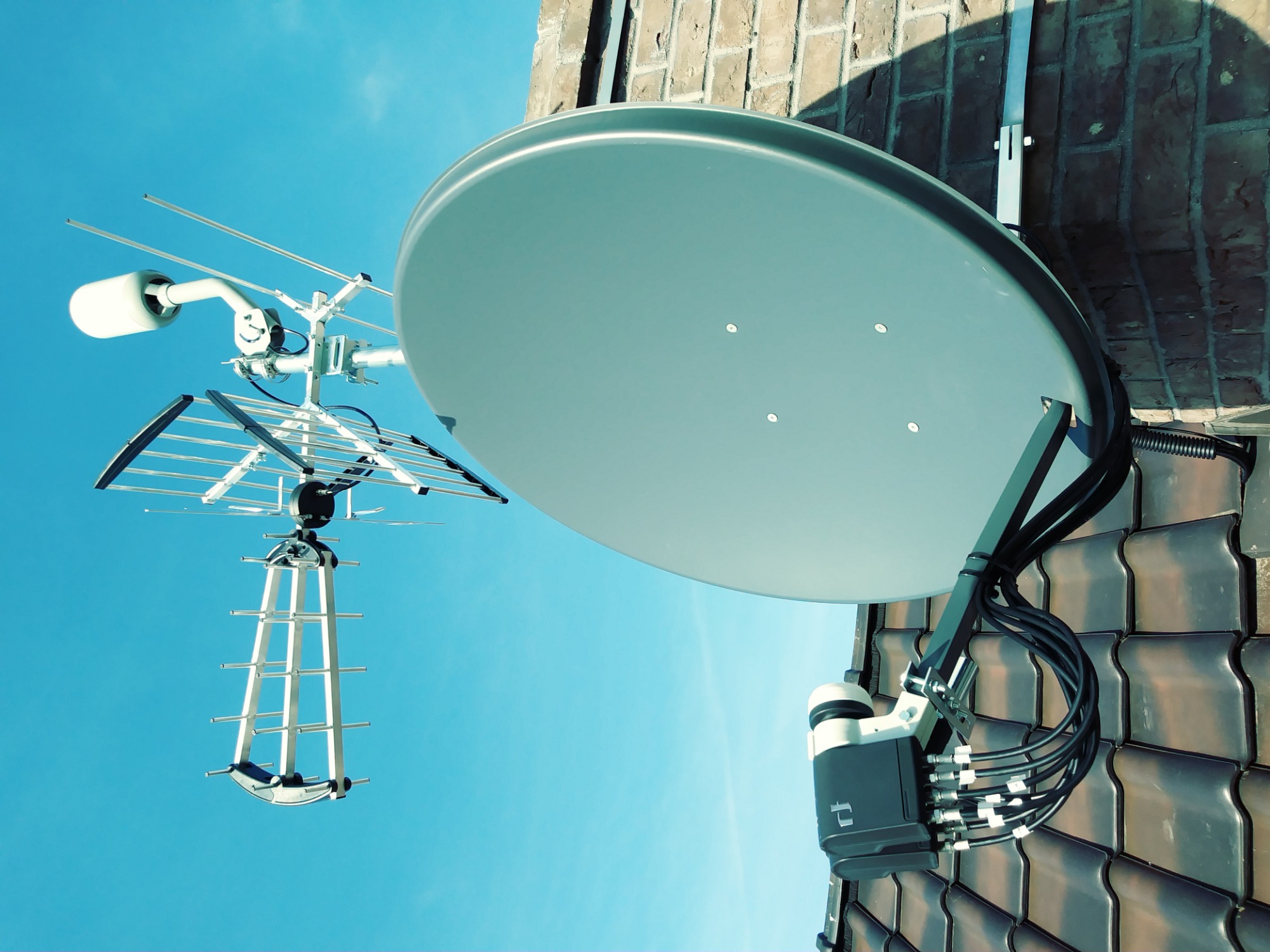 Antena satelitarna i antena naziemna zamontowane na dachu z dachówki ceramicznej, widok z dołu na tle błękitnego nieba.